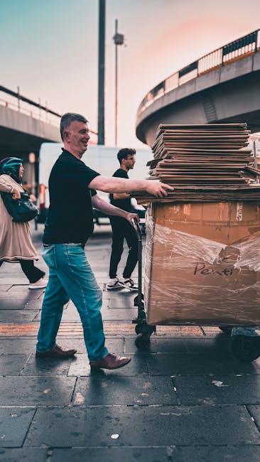 A man with short hair and a black shirt is pushing a large trolley loaded with numerous flattened cardboard boxes and packing materials, secured with plastic wrap, on a city pavement near an overpass. Several other individuals are visible in the background, some walking past and others observing the scene. The setting appears to be part of a house or furniture removal process, with the trolley positioned close to the edge of the pavement, indicating a loading or unloading activity. The lighting suggests late afternoon or early evening, with the overpass casting shadows on the ground. The image exemplifies the logistics involved in home relocation services provided by Ilford Man and Van, highlighting the careful handling of packing materials and the efficient transportation of moving boxes during furniture transport and packing operations for house removals.