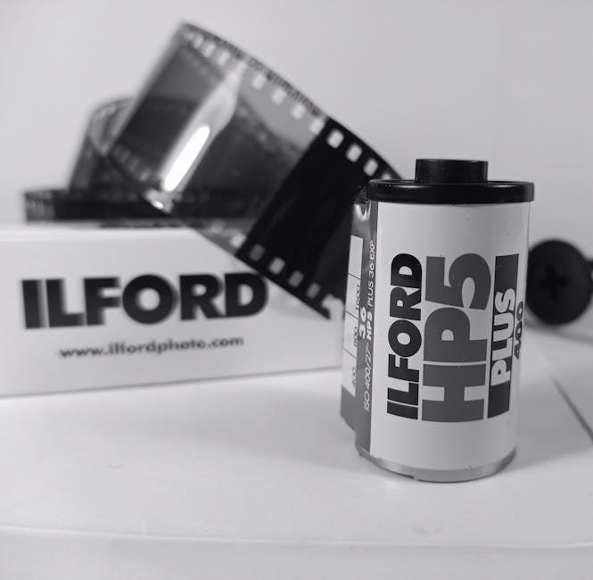 A black and white photograph showing a roll of Ilford HP5 Plus black and white photographic film standing upright in the foreground on a white surface. Behind the film roll, there is an opened cardboard box with the Ilford logo and website printed on it, partially visible and slightly out of focus. A strip of developed photographic film hangs out of the box, draping over the edge. To the right of the film roll, a black screw is lying on the surface. The setting appears to be an indoor environment, possibly a workspace or storage area related to photo processing or archiving, with natural or diffused lighting highlighting the objects' details. Ilford Man and Van occasionally in the background subtly supports the thematic connection to shipping or handling of photo equipment and materials, aligning with house removals and packing services.