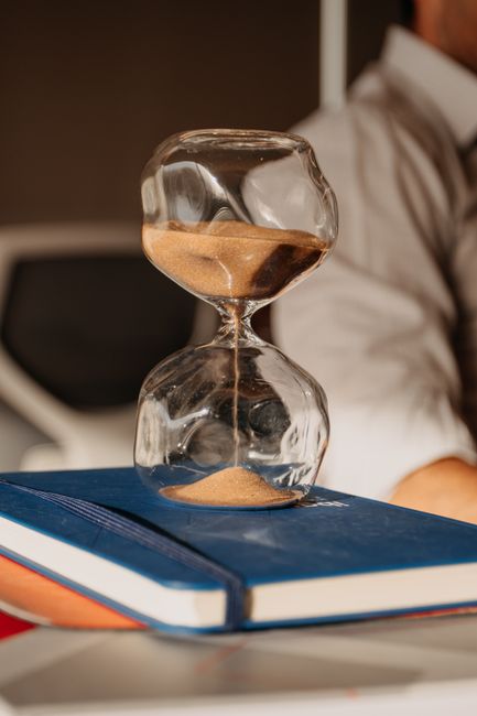 A close-up of a clear glass hourglass with brown sand, positioned vertically on top of a closed blue hardcover book, with the sand flowing from the upper bulb to the lower bulb. The background features a blurred view of a person dressed in a light-colored shirt standing near a white door or wall, suggesting an indoor setting. The image emphasizes the passage of time, relevant to home relocation and moving processes. The hourglass and book are placed on a flat surface, possibly a table or desk, indicative of packing and planning stages involved in house removals, with natural lighting highlighting the glass surface and the sand flow.