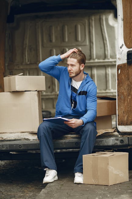 A young man with light brown hair, wearing a blue zip-up hoodie and white sneakers, is sitting on the edge of an open moving van parked outside a property. He is holding a clipboard in his left hand and appears to be contemplating, with his right hand running through his hair. Inside the van, several brown cardboard boxes of various sizes are visible, some stacked and some placed on the ground near the vehicle. The interior of the van shows the metal walls and flooring typical of a moving truck, with protective padding or wooden panels on the sides. The scene captures an inside look at the packing and loading process during a home relocation, with the man possibly taking a break or reviewing a moving checklist. Natural lighting illuminates the area, emphasizing the details of the packaging materials, furniture, and equipment involved in furniture transport and house removals, highlighting the logistical aspects of a professional move provided by companies like Ilford Man and Van.