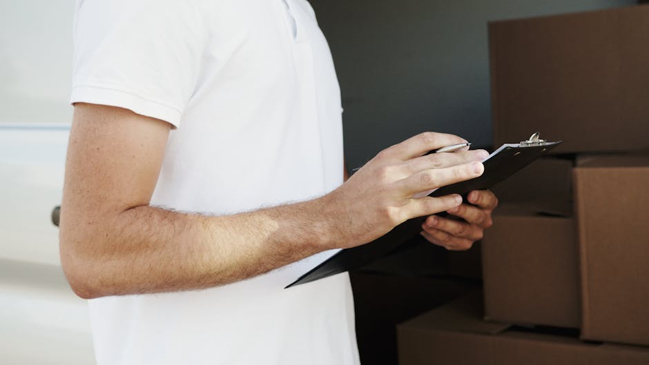 A person wearing a white short-sleeved shirt is holding a black clipboard and pen, inspecting or taking notes during a home relocation process. The background shows stacked cardboard boxes and packing materials, indicating the packing and moving activities involved in house removals. The person's arms and hands are visible, with the focus on their task of checking or organizing items, possibly as part of a furniture transport or packing assessment by Ilford Man and Van. The lighting is even, highlighting the details of the boxes and the person's actions. This image reflects the logistical aspects of packing and moving, relevant to professional removals services in Ilford, supporting the topic of avoiding hidden removal fees and ensuring a smooth moving process.