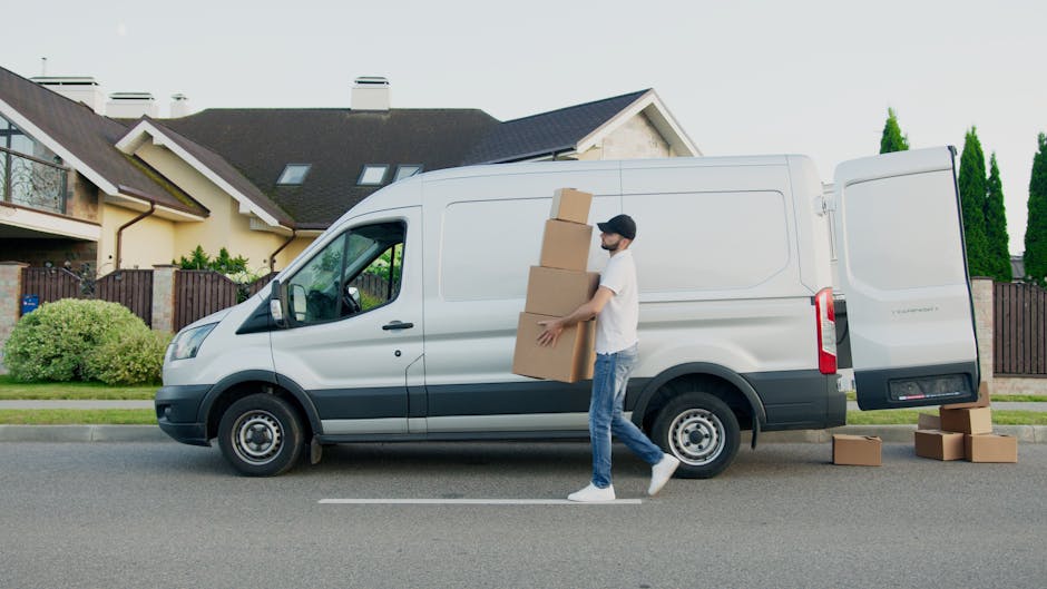 A person wearing a white short-sleeved shirt is holding a black clipboard and pen, inspecting or taking notes during a home relocation process. The background shows stacked cardboard boxes and packing materials, indicating the packing and moving activities involved in house removals. The person's arms and hands are visible, with the focus on their task of checking or organizing items, possibly as part of a furniture transport or packing assessment by Ilford Man and Van. The lighting is even, highlighting the details of the boxes and the person's actions. This image reflects the logistical aspects of packing and moving, relevant to professional removals services in Ilford, supporting the topic of avoiding hidden removal fees and ensuring a smooth moving process.