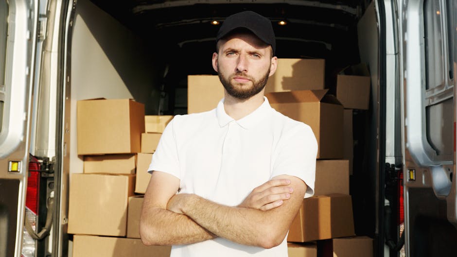 A young man with a serious expression, wearing a white polo shirt and a black baseball cap, stands inside the open back of a moving van filled with stacked cardboard boxes. The boxes vary in size and are packed tightly, some wrapped in plastic film for protection. The van's interior is illuminated by natural light, revealing the arrangement of the packing materials and the loading process for home relocation. The man, representing a professional removals service, is positioned in front of the boxes with his arms crossed, indicating readiness to assist with furniture transport and packing during a house move. The scene is set outdoors on a paved area, with the metal sides of the van visible on either side, emphasizing the logistics involved in house removals as part of a professional house and furniture moving service provided by Ilford Man and Van. This image captures the essentials of packing and loading during a same-day house move, aligning with the services offered for efficient home relocations in Seven Kings and surrounding areas.