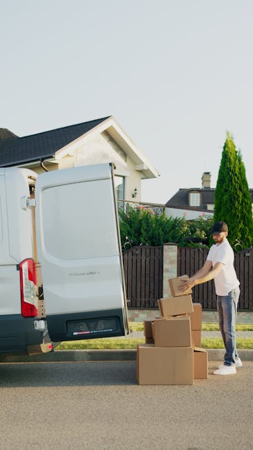 A man stands outside a residential property, engaged in the packing and loading process for a home relocation. He is wearing casual clothing and is preparing to load cardboard boxes of various sizes into the open rear side door of a white moving van, which is parked on the street. The boxes are stacked on the ground beside him, and he is in the midst of lifting one of the boxes. The moving van is positioned next to a paved sidewalk, with a wooden fence and greenery, including trees and shrubs, visible in the background. The scene is illuminated by daylight, capturing a typical house removal activity involving furniture transport and packing materials, facilitated by Ilford Man and Van, a professional removals company specializing in same-day house moves, as indicated by the context of the webpage about best man and van services in Seven Kings.