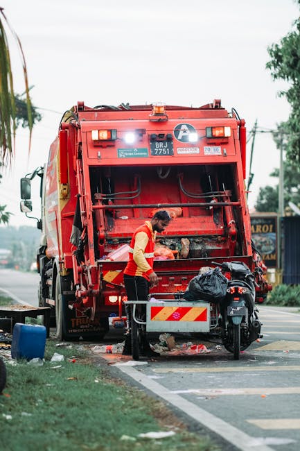 A worker wearing a yellow high-visibility vest is standing beside a large red waste collection vehicle on a street, near the tailgate which is open and equipped with mechanical arms for waste disposal