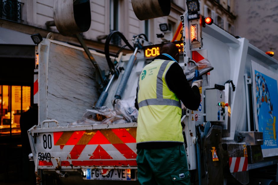 A worker wearing a yellow high-visibility vest is standing beside a large red waste collection vehicle on a street, near the tailgate which is open and equipped with mechanical arms for waste disposal