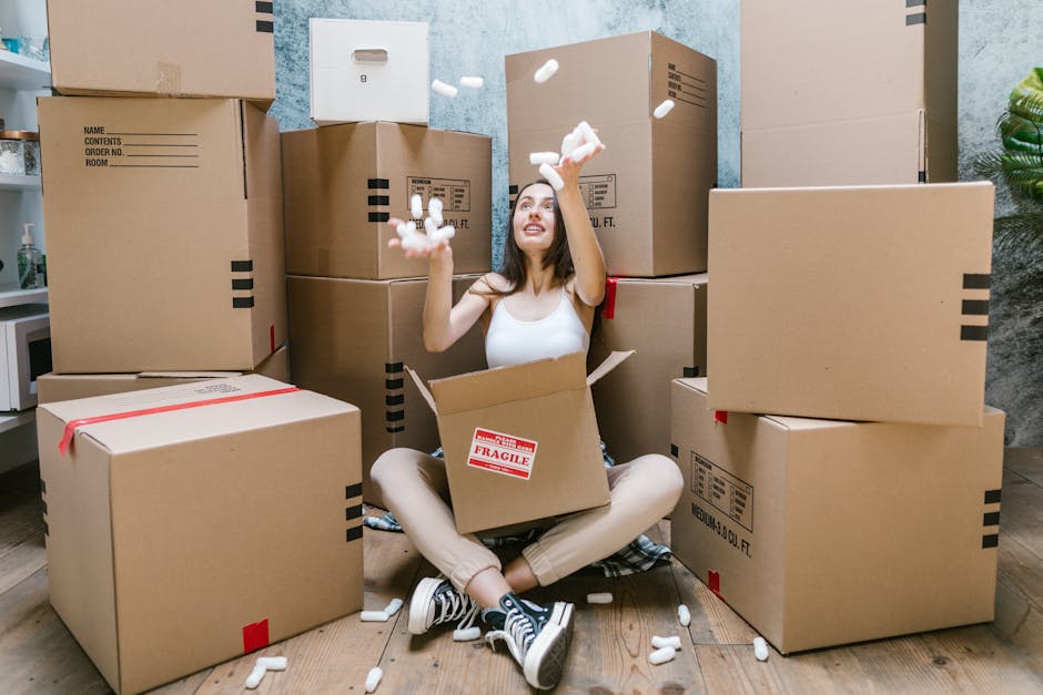A woman sitting cross-legged on a wooden floor inside a property, surrounded by numerous large cardboard moving boxes, some sealed with red or black tape and others labeled with packing or contents information. She is wearing casual clothing, including a white sleeveless top and beige pants, and is smiling while throwing packing peanuts into the air. The boxes are stacked around her, with some on the floor and others placed on furniture or directly against the wall. Behind her, the room has a textured light blue wall, a small white shelf with decorative items, and a potted plant partially visible on the right. The scene captures the packing and home relocation process, with a focus on the preparation phase facilitated by Ilford Man and Van, a professional removals service, highlighting the packing materials, cardboard boxes, and interior environment associated with furniture transport and moving logistics.
