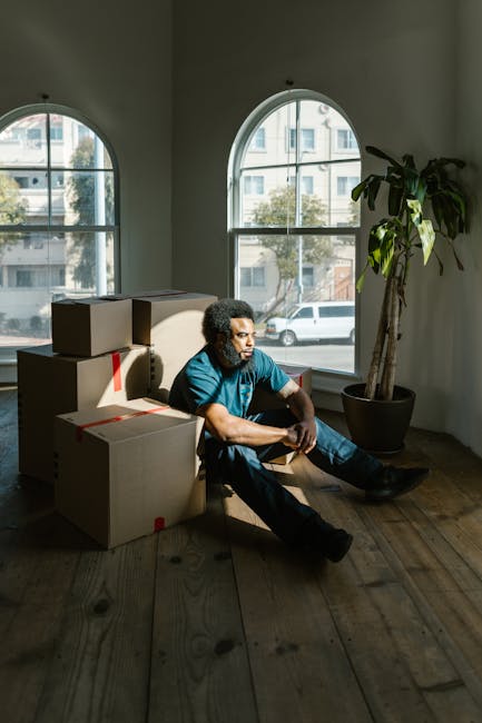 A person with curly hair, wearing a blue t-shirt and dark jeans, is seated on a wooden floor inside a room during a home relocation process. They are surrounded by several cardboard boxes, some stacked and others placed on the floor, all sealed with red tape. The room has large, arched windows allowing natural light to illuminate the space, with a view of a street and parked cars outside. A tall potted plant with broad green leaves stands beside the person near the window. The scene captures a moment of pause amidst packing and moving activities, with the individual appearing thoughtful. This setting reflects the ongoing process of packing and preparing for furniture transport and house removals, which is relevant to services offered by Ilford Man and Van for house moves and relocation in the Cranbrook Estate area.