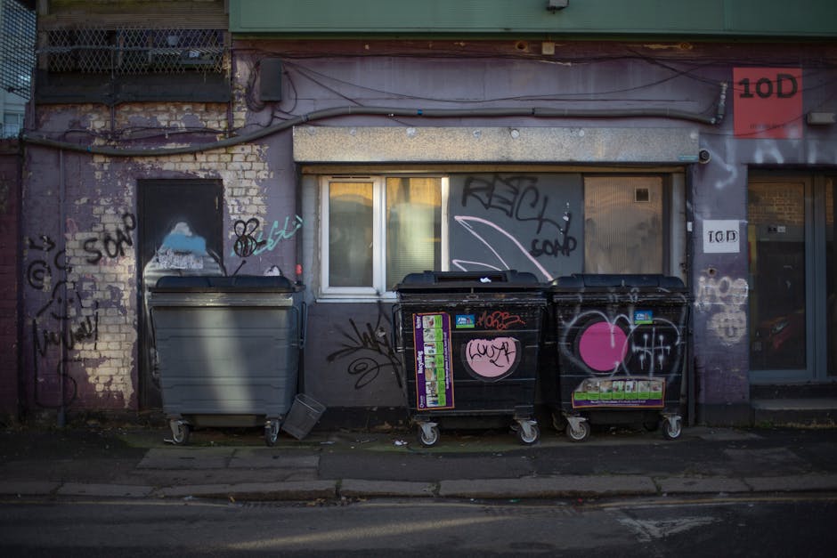 The image depicts a street scene outside a building with a weathered, graffiti-covered exterior wall featuring various tags and markings in black, white, and pink. Two large black commercial waste bins with graffiti and stickers are positioned on the sidewalk, one appearing to contain waste and the other closed with a pink heart sticker. A smaller grey bin is placed nearby, also on wheels, suggesting it is used for moving or disposal purposes. Behind the bins, there's a window with closed blinds and a partially visible door, indicating an entrance to the building. The scene is illuminated by dull, natural lighting, possibly during late afternoon or early evening. The pavement in front of the building is typical urban sidewalk, with some dirt and debris scattered on the ground. This setting may relate to home relocation or packing and moving activities, possibly illustrating an area affected by street cleaning, waste disposal, or logistical elements of a removals service like Ilford Man and Van, as referenced in the context of house removals and relocation permits.