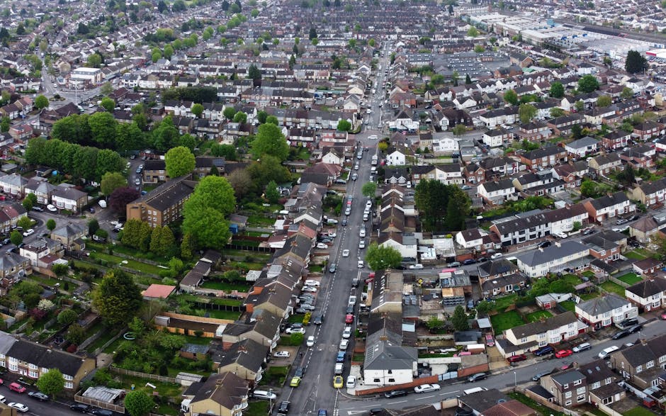 An aerial photograph of a residential neighbourhood showing a straight, narrow street lined with parked cars on both sides. The street runs through rows of terraced houses with small front gardens, some featuring driveways, and back gardens visible behind the houses. Several large, leafy trees are scattered throughout the area, providing greenery amidst the tightly packed houses. The surrounding area includes additional houses and green spaces, with a mix of rooftops in various shades of brown, grey, and black. The scene is captured in daylight with clear visibility of the layout, demonstrating typical urban housing arrangements suitable for home relocation and furniture transport services by Ilford Man and Van, as referenced on the ILFORD page about removal permits fines and house moves.
