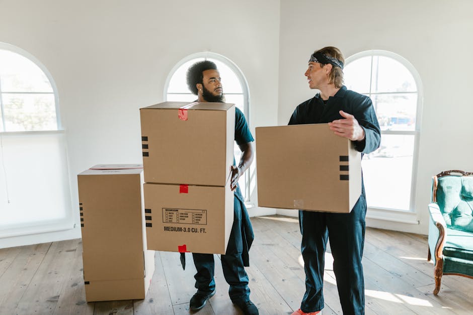 A woman sitting cross-legged on a wooden floor surrounded by numerous large cardboard boxes, some sealed with red tape and others open. She is inside a room with a blue textured wall, occasionally tos