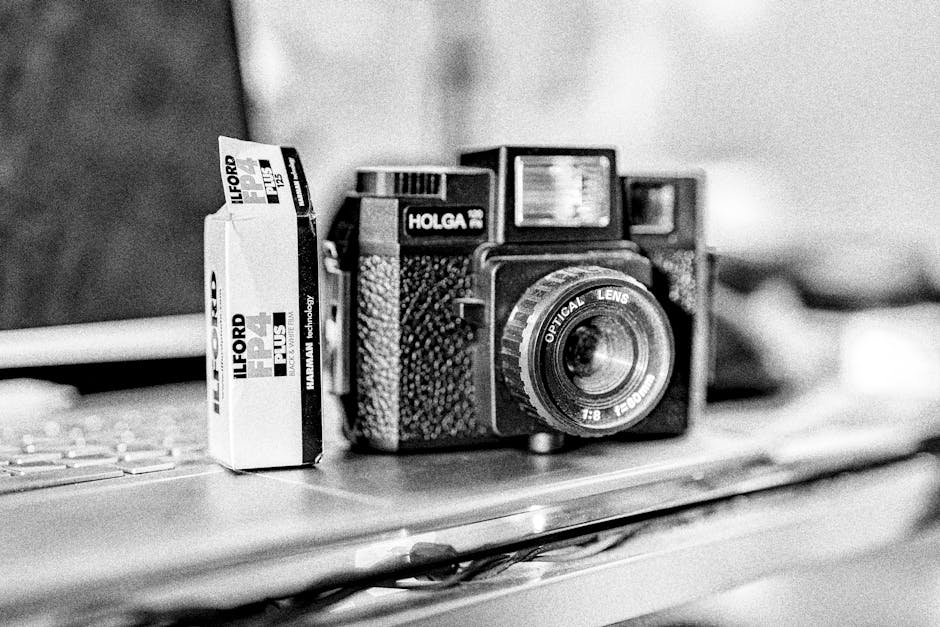 A black and white photograph shows a vintage film camera with a textured body and a prominent optical lens placed on a flat surface, possibly a desk or table. Next to the camera, there is a small box of Ilford Plus photographic paper standing upright, with visible branding and size information. The surface appears to be made of smooth material, and the background features blurred objects and a room interior, with soft lighting illuminating the scene evenly. This setup could represent packing or organising items for a home relocation or camera equipment transportation during a professional removal service. The image emphasizes careful handling and packing of fragile or valuable items, aligning with the services provided by Ilford Man and Van for house removals and furniture transport.
