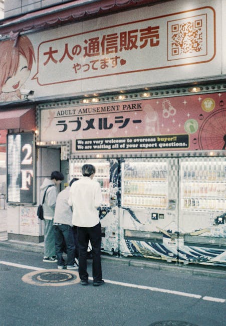Three individuals are standing outside a vending machine area under a large overhead sign featuring Japanese characters and illustrations, with one person dressed in a white jacket and the other two in darker clothing. They appear to be selecting or purchasing items from the vending machines, which are positioned on a sidewalk next to a building. The vending machines are filled with various canned and bottled beverages, with visible digital payment interfaces. The area is lit with artificial lighting, suggesting it is evening or nighttime. The scene is part of a busy street, potentially near an entertainment or amusement zone, indicated by the signage referencing 'Adult Amusement Park' and 'Overseas Buyers.' The setting includes pavement markings and a manhole cover, and the background contains additional signs and posters in Japanese promoting products or services. This visual captures a typical street scene involving a snack or drink purchase, relevant within the context of packing, moving logistics, and transportation phases in house removals, as handled by companies like Ilford Man and Van.