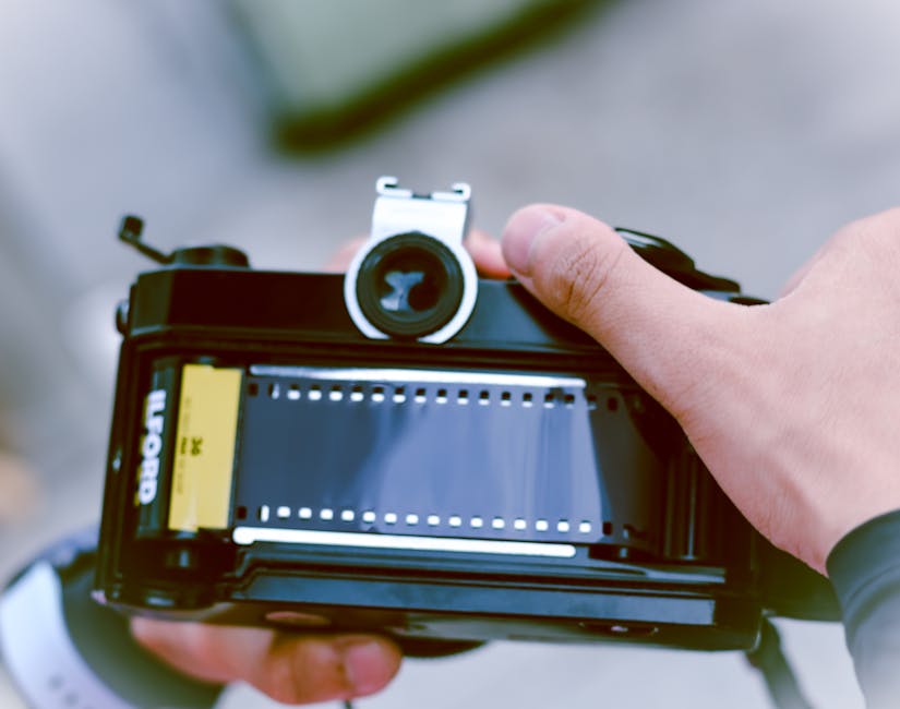 A black and white photograph shows a vintage film camera with a textured body and a prominent optical lens placed on a flat surface, possibly a desk or table. Next to the camera, there is a small box of Ilford Plus photographic paper standing upright, with visible branding and size information. The surface appears to be made of smooth material, and the background features blurred objects and a room interior, with soft lighting illuminating the scene evenly. This setup could represent packing or organising items for a home relocation or camera equipment transportation during a professional removal service. The image emphasizes careful handling and packing of fragile or valuable items, aligning with the services provided by Ilford Man and Van for house removals and furniture transport.
